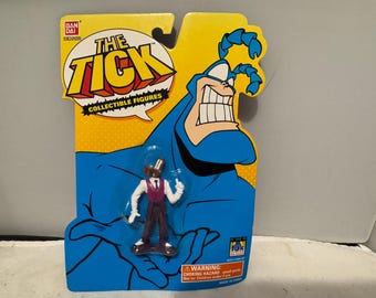 May include: A collectible action figure of a character from "The Tick" cartoon series. The figure is on a yellow and blue backing card with the show's logo. The figure is of a man in a suit and the card has a warning about small parts.
