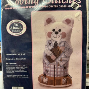 May include: A counted cross stitch kit featuring a white bear wearing a blue and white plaid shirt and brown shoes. The kit is titled "Barney Bear" and is part of the "Sit Abouts Collection". The finished size is 10 inches by 15 inches.