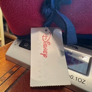 K&ouml;nnte beinhalten: Ein gerollter, roter und blauer Artikel mit einem blauen Band und einem wei&szlig;en Etikett mit der Aufschrift "Disney Store Exclusive" in Rot. Der Artikel liegt auf einer silbernen Waage, das Gewicht betr&auml;gt 2,8 Gramm.