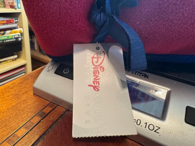 K&ouml;nnte beinhalten: Ein gerollter, roter und blauer Artikel mit einem blauen Band und einem wei&szlig;en Etikett mit der Aufschrift "Disney Store Exclusive" in Rot. Der Artikel liegt auf einer silbernen Waage, das Gewicht betr&auml;gt 2,8 Gramm.