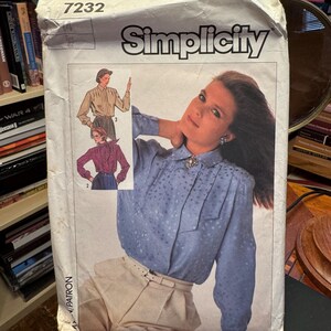 May include: A Simplicity 7232 sewing pattern envelope featuring a light blue long-sleeved blouse with a subtle floral pattern, a collar, and a decorative brooch. The envelope displays illustrations of two blouse variations in tan and maroon.