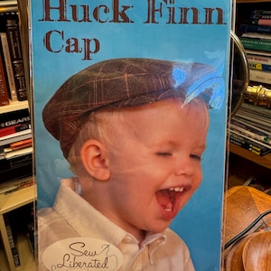 Può includere: Una busta di cartamodello per un berretto Huck Finn Cap, n. 110, di Sew Liberated. La copertina presenta una fotografia di un bambino che indossa un berretto piatto a quadri marroni, con il testo Huck Finn Cap e Sew Liberated su sfondo blu.