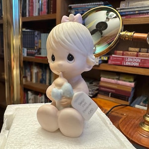 Könnte beinhalten: Eine Biskuitporzellanfigur eines Babys, das eine Flasche mit einer blauen Schleife hält. Die Figur hat eine rosa Schleife auf dem Kopf und sitzt. Ein Etikett lautet "Baby's First Christmas 1985."