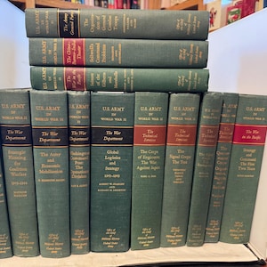 Può includere: Una collezione di tredici libri con copertina rigida verde intitolati U.S. Army in World War II, disposti su due file. I dorsi presentano bande orizzontali nere e rosse con scritte dorate che descrivono specifici argomenti di storia militare.