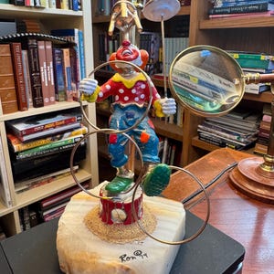 May include: A colorful clown figurine balancing hoops, a plate, and a magnifying glass. The clown wears a red polka-dot shirt, blue pants, and green shoes. The figurine is mounted on a marble base with the artist's signature.