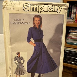 Può includere: Un modello di cucito Simplicity vintage con una donna in un lungo abito abbottonato con cintura nera. Il modello è etichettato "Cathy Hardwick" e include informazioni sulle taglie. La confezione è bianco sporco con testo nero e blu.