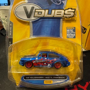 May include: A blue 1965 Volkswagen 1600 TL Fastback die-cast toy car with red and white flames and stars on the side. The car is part of the V-Dubs series and is labeled as #048.