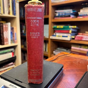 以下が含まれることがあります： 銀色の文字で「International Cook Book」と書かれた、ヴィンテージの赤い料理本。 縦に立っており、「World's Famous Chefs」と「The Red White Stores」の文字が書かれています。 黒い計量器の上に置かれ、背景には本棚があります。