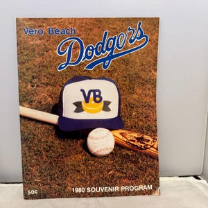 Może przedstawiać: Okładka programu pamiątkowego z drużyną baseballową Vero Beach Dodgers. Okładka przedstawia nazwę drużyny w niebieskim skrypcie, czapkę baseballową, piłkę i kij. Program pochodzi z 1980 roku.