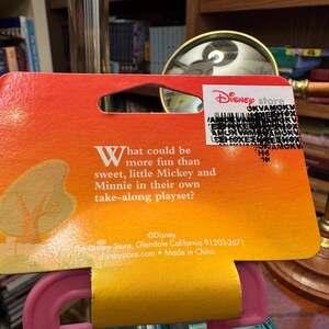 May include: A Disney Store product tag with a gradient orange and yellow background. The tag features text asking "What could be more fun than sweet, little Mickey and Minnie in their own take-along playset?" A Disney Store logo is visible.