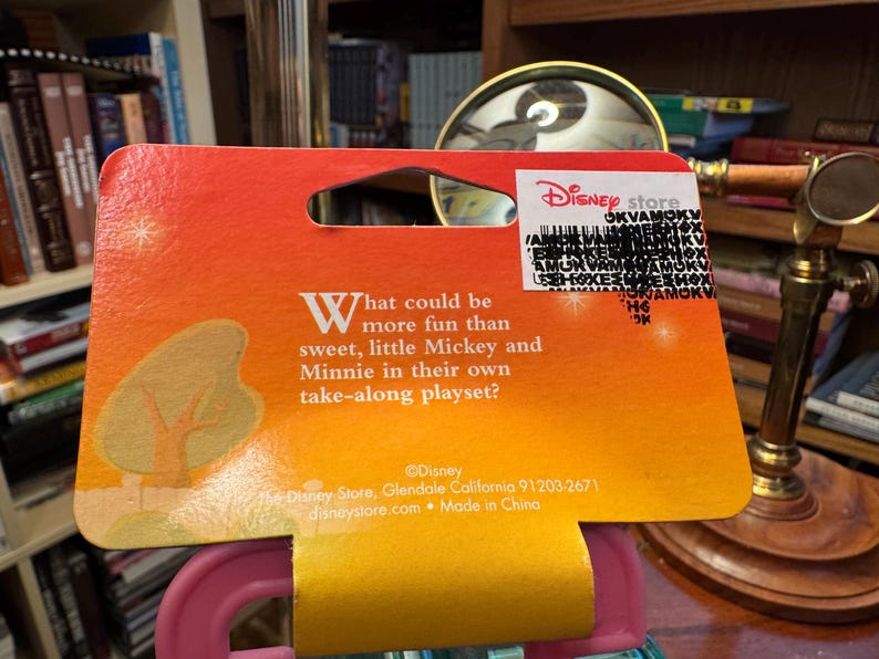 May include: A Disney Store product tag with a gradient orange and yellow background. The tag features text asking "What could be more fun than sweet, little Mickey and Minnie in their own take-along playset?" A Disney Store logo is visible.