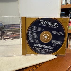 May include: A Chesky Records CD in a gold-colored case. The CD is dark blue with gold text and a gold center. The case is open, revealing the CD and its tracklist. The text "Superior Gold" is visible.