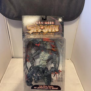 Może przedstawiać: Figurka akcji Ultra Spawn Dark Ages w oryginalnym opakowaniu. Figurka to szczegółowe, ciemne stworzenie z czerwonymi akcentami i skrzydłami. Opakowanie zawiera logo Spawn i tekst "The Epic Saga of America's #1 Hero!"