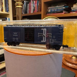 May include: A navy blue model train boxcar with white lettering and a logo, encased in a clear plastic display case. The boxcar has black wheels and is set on a round, light-colored surface. The background includes bookshelves.