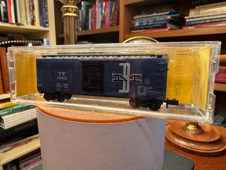 May include: A navy blue model train boxcar with white lettering and a logo, encased in a clear plastic display case. The boxcar has black wheels and is set on a round, light-colored surface. The background includes bookshelves.