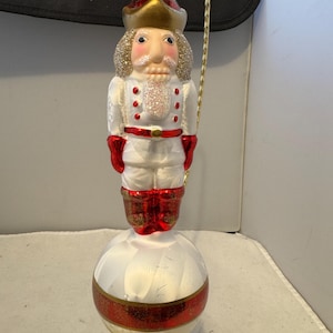 Puede incluir: Adorno navideño decorativo con forma de soldado cascanueces. El adorno presenta un uniforme blanco con detalles rojos, un sombrero dorado y una cara brillante. Está montado sobre una bola a rayas blancas y rojas.