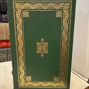 May include: A green hardcover book with gold decorative detailing. The cover has a gold border and a central gold emblem. The book is part of a collection of books.