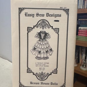 May include: A vintage sewing pattern for a Shirley costume, inspired by "The Little Colonel." The cover features a detailed illustration of the dress, framed by ornate black borders. The text reads "Easy Sew Designs" and "Brown House Dolls."