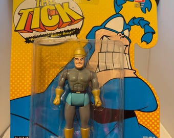 May include: The Tick action figure in original packaging. The figure is gray, blue, and yellow, with a helmet and gloves. The packaging is yellow and blue with the words "The Tick" and "Human Bullet". The packaging also has the Fox Kids Network logo.