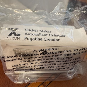 May include: A clear plastic bag containing a sticker maker. The label reads "Sticker Maker" in English, French, and Spanish. A warning about suffocation from plastic bags is also printed on the label.