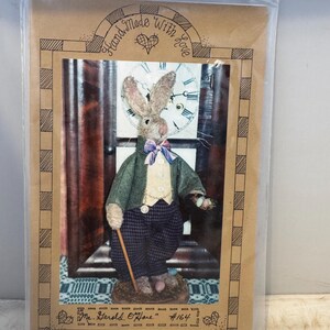 May include: A pattern for a handmade plush toy rabbit wearing a green jacket, plaid pants, a bow tie, and holding a walking stick. The pattern is titled "Mr. Gerald O'Hare" #164 and is hand-made with love.