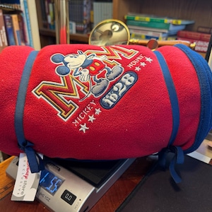 K&ouml;nnte beinhalten: Eine gerollte rote Fleece-Decke mit Mickey Mouse-Stickerei, blauen Verzierungen und Bindeb&auml;ndern. Die Decke zeigt "Mickey Mouse 1928" und liegt auf einer Digitalwaage. Ein Disney Store-Etikett ist sichtbar.