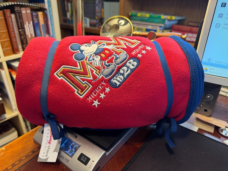 K&ouml;nnte beinhalten: Eine gerollte rote Fleece-Decke mit Mickey Mouse-Stickerei, blauen Verzierungen und Bindeb&auml;ndern. Die Decke zeigt "Mickey Mouse 1928" und liegt auf einer Digitalwaage. Ein Disney Store-Etikett ist sichtbar.