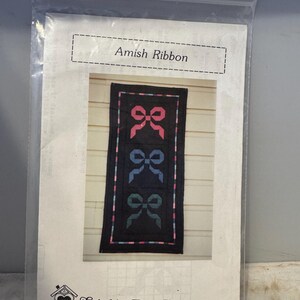 May include: A quilting pattern for a quilt with a black background and three rows of colorful bows. The pattern is titled "Amish Ribbon" and is from "Stitch'n Post Patterns".