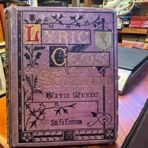 May include: A vintage book titled "Lyric Gems of Scotland With Music" in a purple cover with gold lettering. The cover features decorative floral and banner designs. The book is an antique, likely from the Sol Fa Edition.
