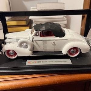 Op de afbeelding: Witte 1938 Buick Century Convertible Coupé modellauto in een vitrine met een zwart dak en een rood interieur. De auto staat op een zwarte basis met een zilveren plaquette waarop "1938 Buick Century Convertible Coupé" staat.
