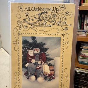 May include: A craft pattern titled "All Gathered Up" featuring a snowman in a winter scene. The snowman wears a red coat and holds a sign. The pattern is in a clear plastic sleeve.
