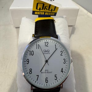 May include: A silver and black Q&Q Quartz watch with a white face and black leather band. The watch face has black numbers and hands, and the words "Q&Q Quartz" and "IPS Water Resist" are printed on the face.