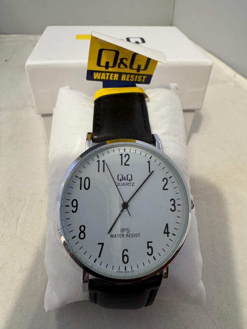 May include: A silver and black Q&Q Quartz watch with a white face and black leather band. The watch face has black numbers and hands, and the words "Q&Q Quartz" and "IPS Water Resist" are printed on the face.