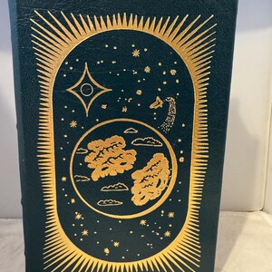 Può includere: Una copertina di libro verde scuro con un design dorato. Il design presenta un'immagine stilizzata di un pianeta con nuvole, una stella cadente e una stella con un cerchio al centro. Il design è circondato da un cerchio di stelle dorate.