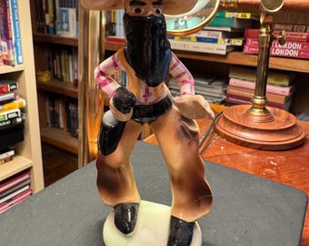 May include: A ceramic cowboy figurine with a large hat, black beard, and a holster. The figurine is wearing a pink and white striped shirt, a yellow vest, and brown trousers. The base is oval-shaped and painted with light green and white colours.