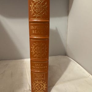 Può includere: Un libro rilegato in pelle marrone con lettere dorate. Il titolo è "Infirmary Beach" di Jack McDevitt. Il libro è una prima edizione firmata.