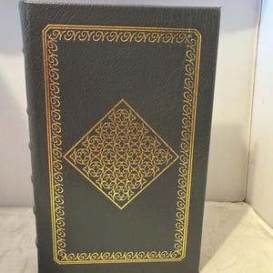 Puede incluir: Un libro encuadernado en cuero verde oscuro con un diseño en relieve dorado en la portada. El diseño presenta una forma de diamante con intrincados patrones florales.