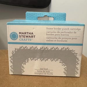 Puede incluir: Una caja de cartucho de perforación de bordes de marco Martha Stewart Crafts. La caja es azul claro y blanca con un diseño de brote de hoja. El texto en la caja incluye "cartucho de perforador" y "brote de hoja".