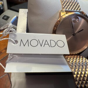 May include: A rose gold Movado watch with a textured band and a minimalist face. The watch is displayed on a white cushion inside a box, with a tag that reads "MOVADO".