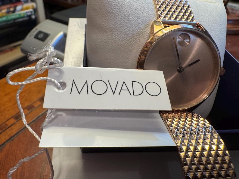 May include: A rose gold Movado watch with a textured band and a minimalist face. The watch is displayed on a white cushion inside a box, with a tag that reads "MOVADO".