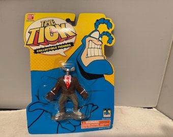 Op de afbeelding: Verzamelbare actiefiguur van The Tick, een superheld, op een gele en blauwe kaart. De figuur draagt een bruin pak, een grijze broek en een rode stropdas. De verpakking bevat de woorden "The Tick" en "Collectible Figures".