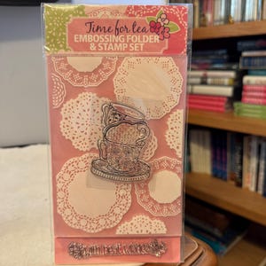 May include: A clear plastic package containing a tea-themed embossing folder and stamp set. The package features a pink background with white doily patterns and the text "Time for tea EMBOSSING FOLDER & STAMP SET". A tea cup stamp is visible.