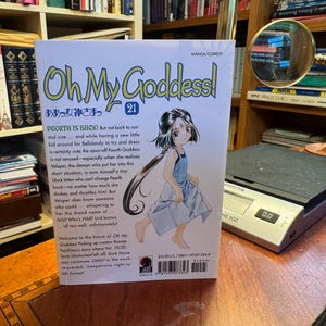 May include: The back cover of the Oh My Goddess! volume 21 manga book features a central illustration of a young girl with long dark hair wearing a light blue dress. The text provides a plot summary, a barcode, and a price of $10.95 U.S.