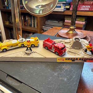 May include: A toy set with miniature emergency vehicles and figures. The set includes a yellow truck, a red fire engine, and two figures. The base is tan with the text "URBAN RESCUE SERIES 1".
