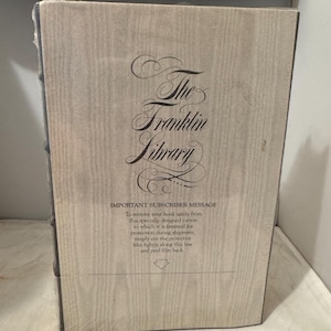 May include: The front cover of a book titled "The Franklin Library". The cover is a light brown with a wood grain texture. The title is written in a cursive font. The text "IMPORTANT SUBSCRIBER MESSAGE" is printed in black below the title. The text below the message explains how to remove the book from its protective packaging.