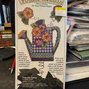 Pode incluir: Um padrão de ponto cruz para uma decoração de planta em vaso. O padrão apresenta um regador xadrez azul e branco com violetas roxas. O padrão chama-se "Potted Whimsy" e custa $ 14,05.