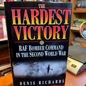 Pode incluir: Um livro intitulado "The Hardest Victory" com capa preta e letras vermelhas e amarelas. O subtítulo é "RAF Bomber Command in the Second World War" de Denis Richards. Uma silhueta de avião é visível.
