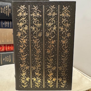 May include: A dark brown book with gold floral designs. The cover features vertical gold floral patterns. Other books are visible in the background. The book's spine is visible, with the title and author's name.