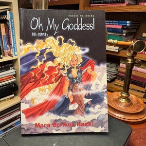 May include: A manga volume titled Oh My Goddess! Mara Strikes Back! by Kosuke Fujishima. The cover features a character with long blonde hair, a dark outfit, and a flowing red cape, posed against a background of dark clouds and swirling mist.