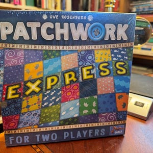 May include: A square board game box titled Patchwork Express for two players by Uwe Rosenberg. The box features a colorful quilt pattern with various fabric designs including fish, flowers, and geometric shapes, with a blue button icon.